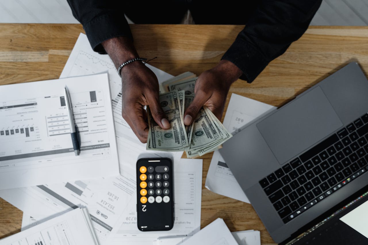 about-us Person counting US dollars, using a calculator and laptop, with financial documents on a wooden desk.